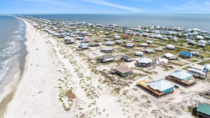 Vista aérea de una zona costera con numerosas casas y edificios frente a la playa, algunos elevados sobre pilotes, a lo largo de una playa arenosa con el océano al fondo.