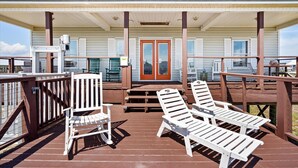 Una terraza techada al aire libre con sillones blancos y una mesa de madera, que conduce a un edificio con puerta roja y contraventanas blancas.