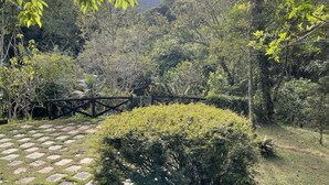 A garden with a paved walkway, a wooden fence, and dense greenery. - Casinha no sítio Calmamente  (Teresópolis)
