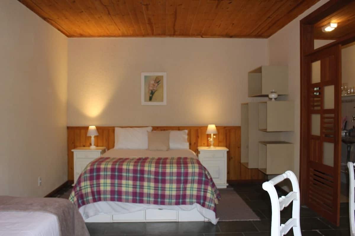 A bedroom with a wooden ceiling, a bed with a plaid blanket, two bedside tables with lamps, a nightstand, and a door leading to another room.