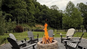 A fire pit with logs, surrounded by Adirondack chairs, set on gravel with a forest backdrop. - Creekfront Cabin • Fire Pit • Fenced Yard • Near Crabtree Falls (Tyro)