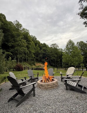 Feuerstelle mit Holz, umgeben von Adirondack-Stühlen, auf Kieselgrund mit Wald im Hintergrund.