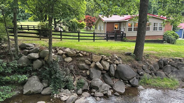 A stone wall, a small stream, and a house with a red roof. - Creekfront Cabin • Fire Pit • Fenced Yard • Near Crabtree Falls (Tyro)