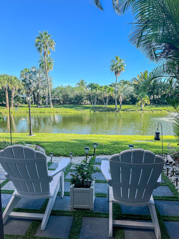Two white Adirondack chairs facing a pond with palm trees and a grassy area in the background.