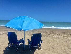 Two blue beach chairs and a blue umbrella on a sandy beach with the ocean in the background.