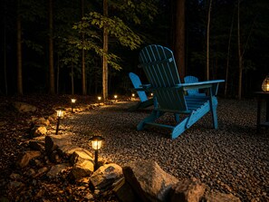 Een bebost gebied met twee Adirondack stoelen en een tafel, omringd door rotsen en kleine lampjes.