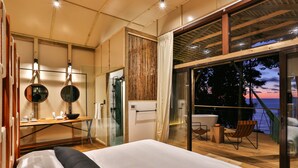 A bedroom with a large bed, a desk with a mirror, and a view of the ocean at sunset. - Corcovado Wilderness Lodge by SCP (Drake Bay)