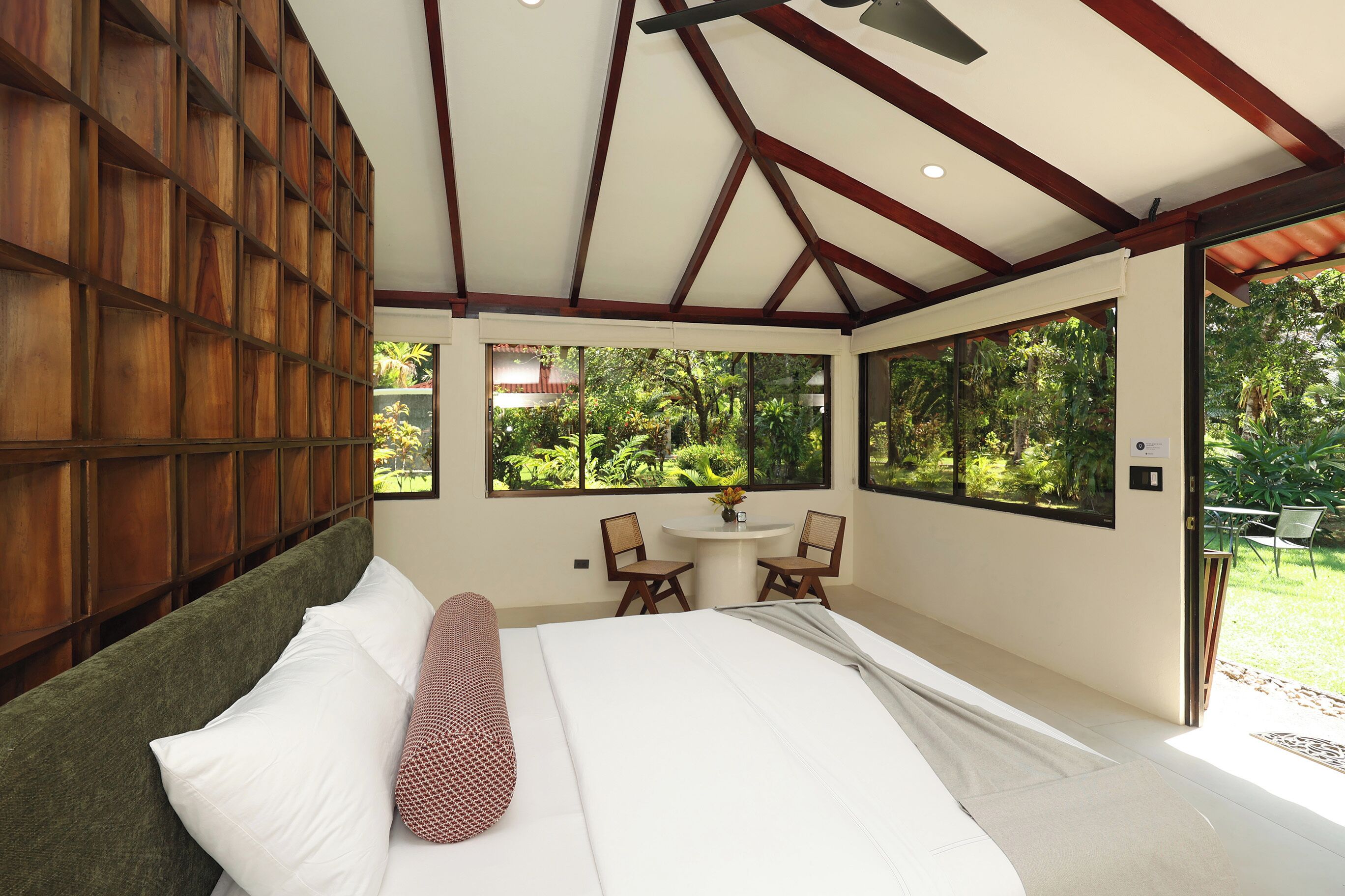 A bedroom with a bed, a wooden headboard, a dining table with chairs, and large windows offering a view of greenery.