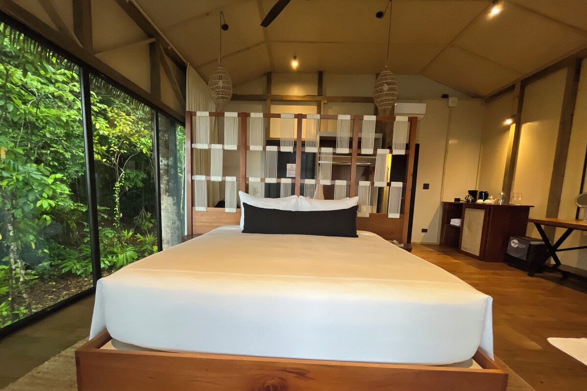 A modern bedroom with a large bed, a wooden headboard, a bedside table, and a view of greenery through large windows.