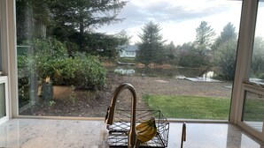 A kitchen with a sink and a window overlooking a backyard with a pond, trees, and a grassy area. - Remote Worker? Retired? Storm Tracker? +Own Canal Dock (Ocean Shores)
