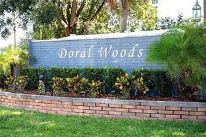 Pared azul com a inscrição "Doral Woods" em letras brancas, delimitada por uma moldura de tijolos e vegetação.