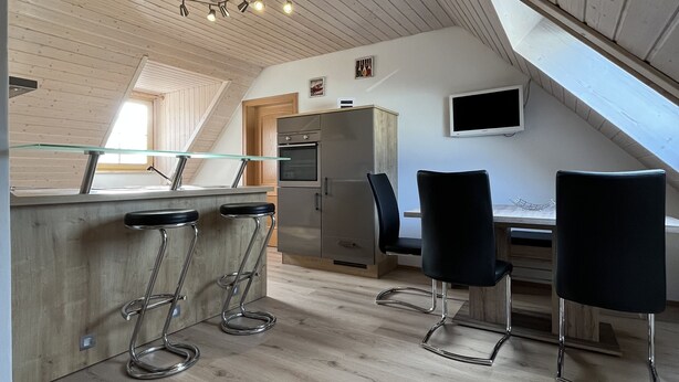 A modern kitchen with wooden ceiling beams, a central island with bar stools, a built-in oven, and a dining area with two black chairs. - Vacation apartment in a quiet location (Schwabmünchen)