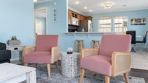 A modern living room with two pink armchairs, a coffee table, and a round side table with a decorative item. A kitchen area is visible in the background with wooden cabinets and a refrigerator. - Great Back to School Dealsl!  4 minute walk to beach! (Bolivar Peninsula)