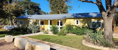 A yellow house with a porch, surrounded by a garden with gravel paths and various plants.