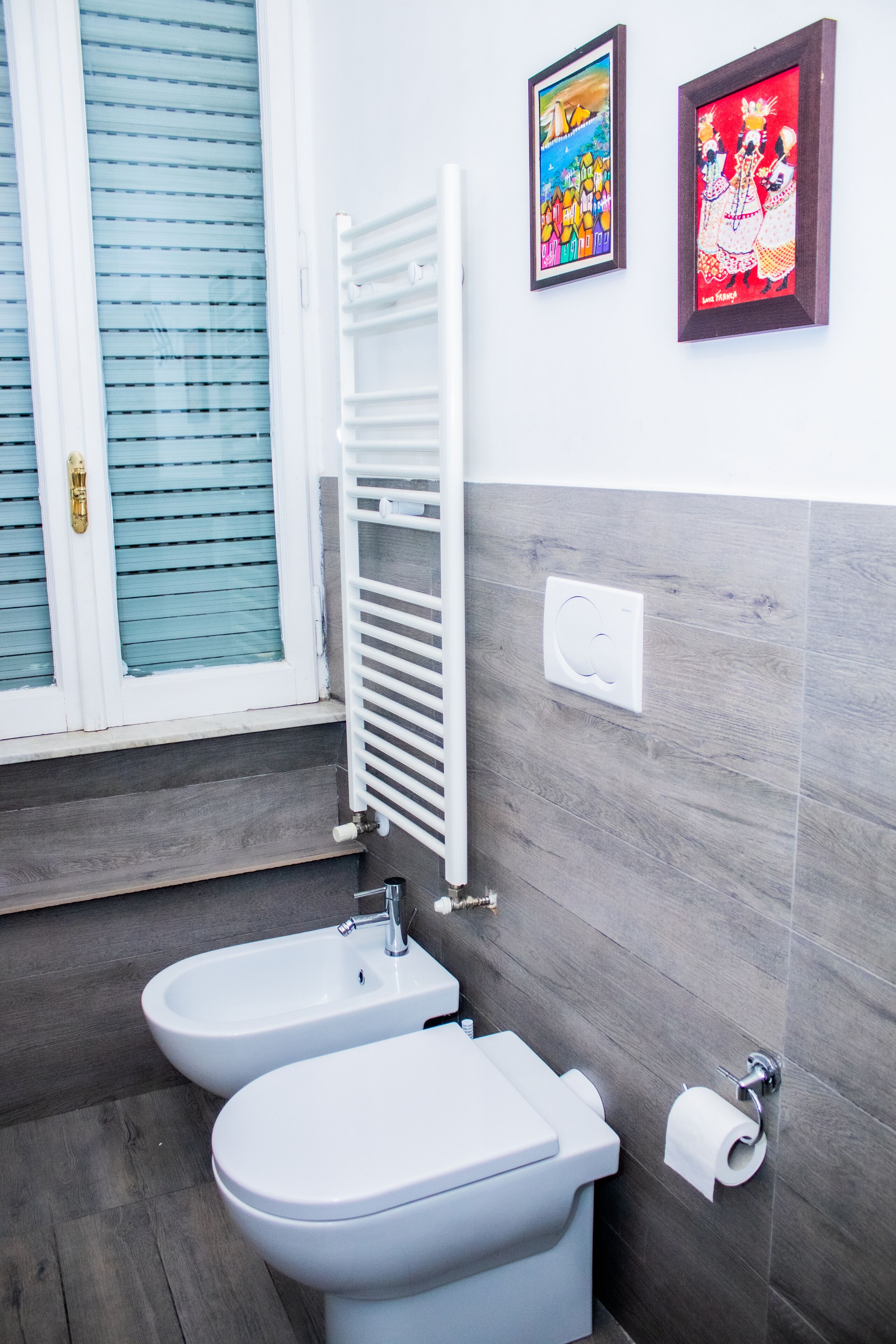 Une salle de bain avec deux toilettes, une fenêtre avec des stores et deux tableaux encadrés au mur.