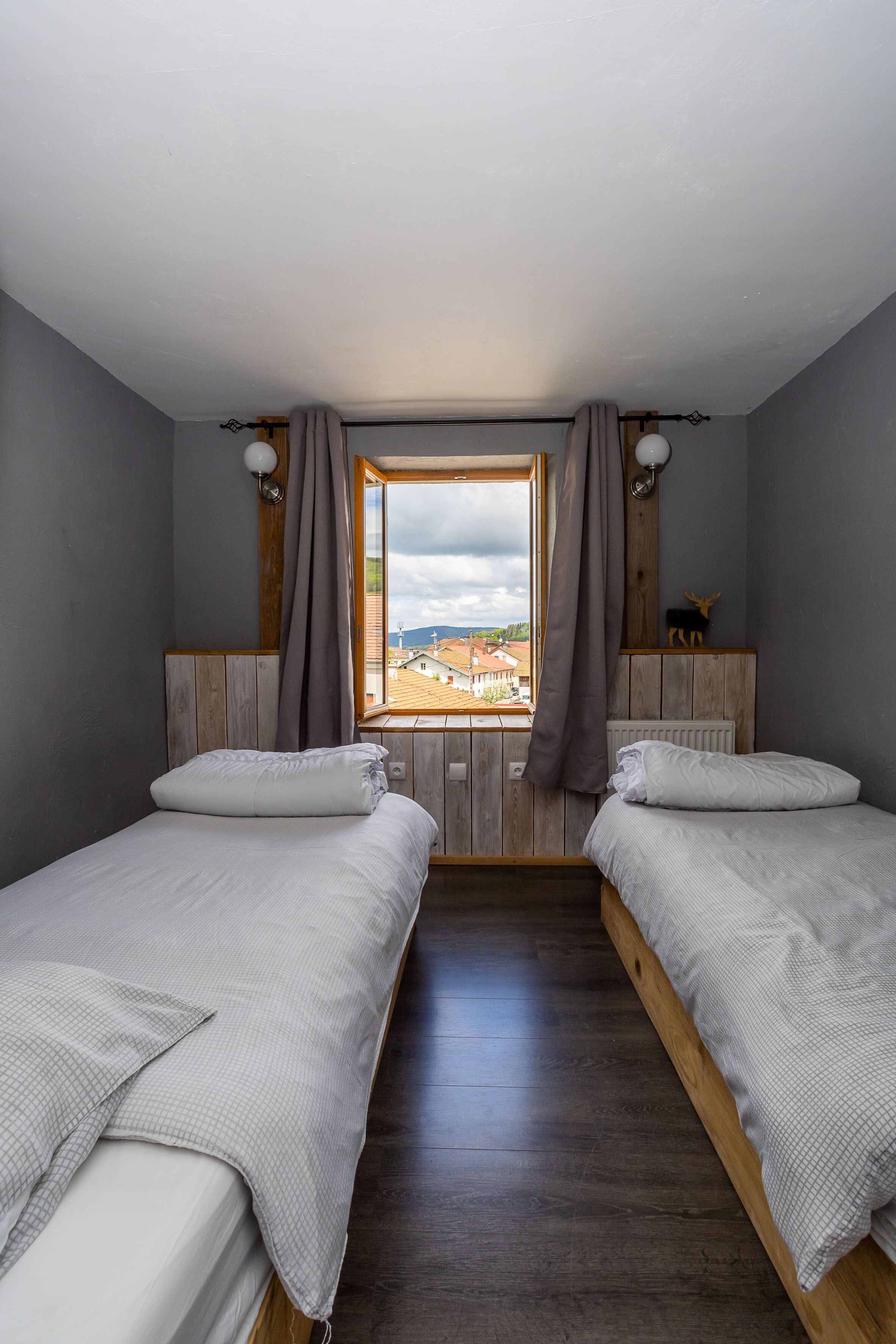 Une chambre avec deux lits, des têtes de lit en bois, une fenêtre donnant sur des immeubles et un luminaire fixé au mur.