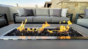 A modern outdoor seating area with a fire feature in the center, surrounded by a stone wall and a grey sofa with cushions. - Private Spa and Balcony Fire Pit With Mountain Views (Washington)