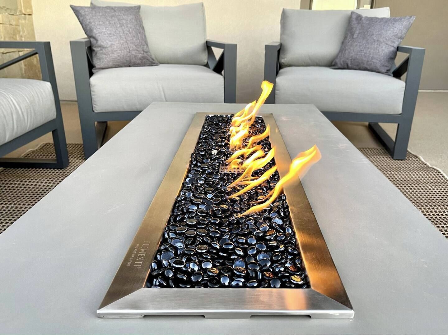 A modern outdoor seating area with a rectangular fire feature surrounded by two grey sofas and matching cushions.