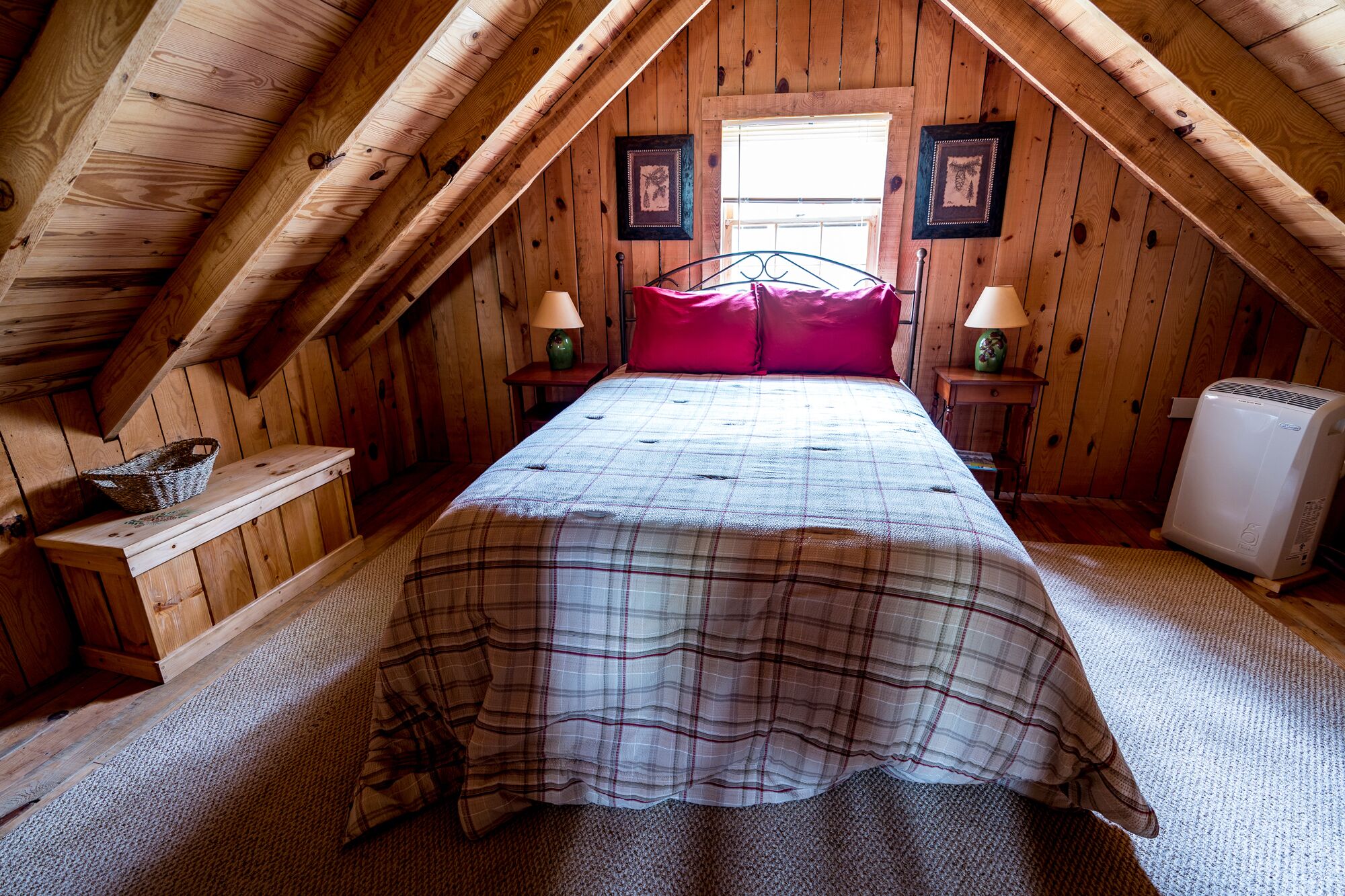 A cozy attic bedroom with a plaid bedspread, two bedside lamps, and a window with wooden shutters.