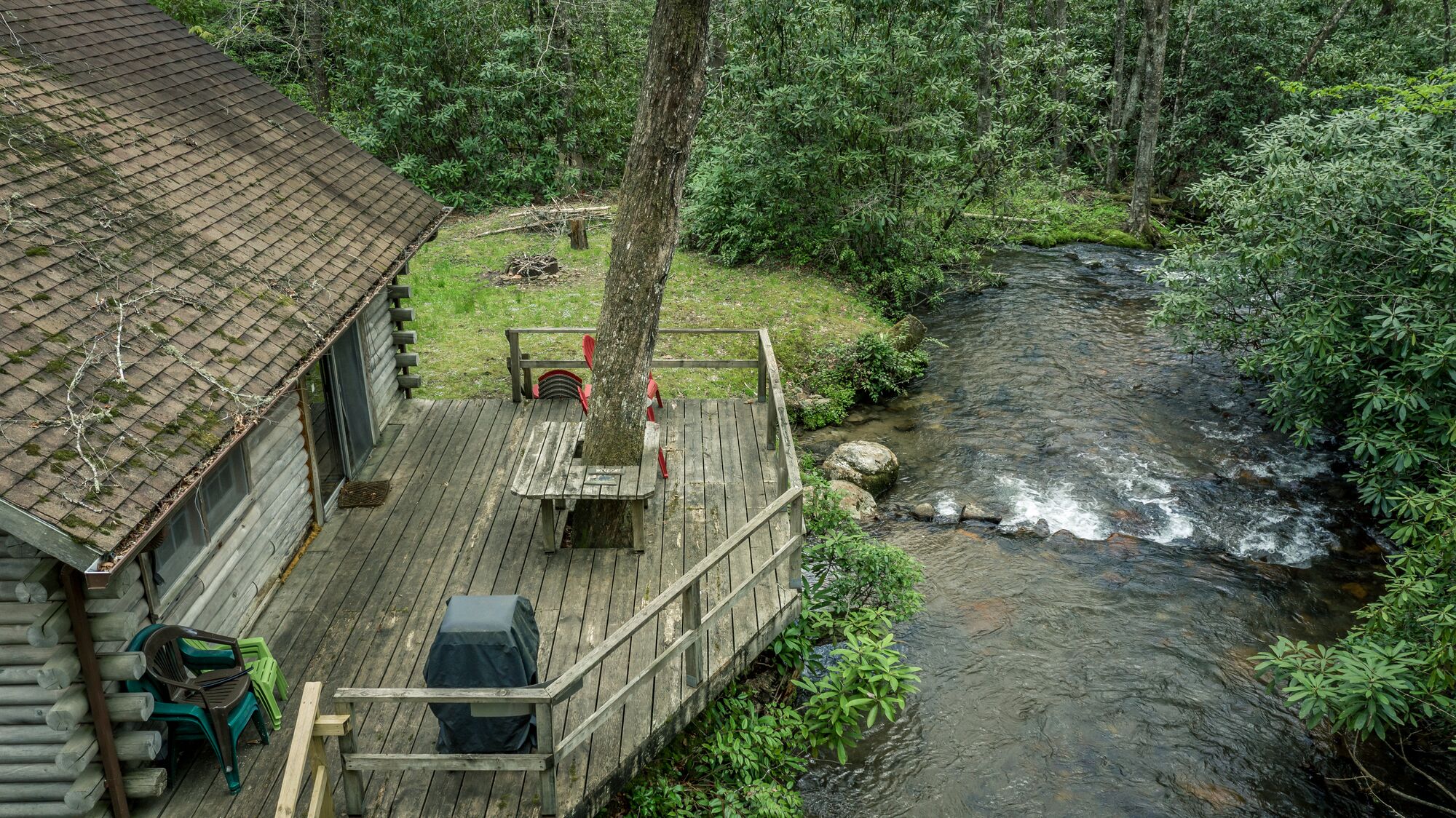 A wooden deck with a table and chairs, overlooking a stream in a forested area.
