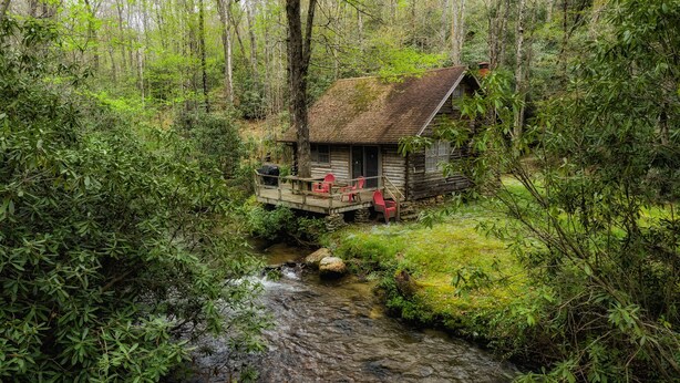 A rustic cabin with a wooden deck and red chairs, surrounded by dense forest and a small stream. - Great Creekside Hideaway Rustic Cabin - Secluded, Creekside Family Cabin (Topton)