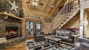 A log cabin interior with a stone fireplace, a wooden staircase, and a cozy seating area with a fireplace rug. - Reeling 'Em Inn by Mountain Oasis | Fightingtown Creekfront (McCaysville)