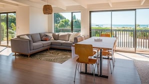 A modern living room with a sectional sofa, a dining table with chairs, and large sliding glass doors leading to a balcony with a view of the ocean.