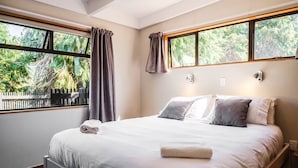A bedroom with a large bed, two windows with curtains, a view of greenery outside, and a small bedside table with a lamp.