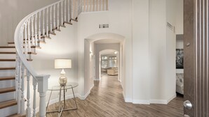 A spacious hallway with a wooden staircase, a glass table with a lamp, and a view into a living area.