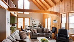 Ein geräumiges Wohnzimmer mit hoher Decke, Holzbalken und großen Fenstern. Es gibt eine Sofagarnitur, einen Couchtisch und einen schwarzen Ledersessel.