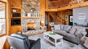 Ein gemütliches Wohnzimmer mit Kamin aus Stein, einer bequemen Couch, einem Fernseher und einer Holztreppe.
