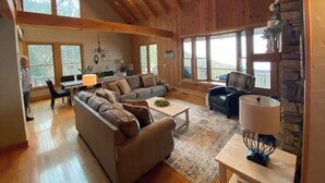 Ein geräumiges Wohnzimmer mit hoher Decke, Holzbalken und großen Fenstern mit Blick ins Freie.