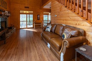 A log cabin interior with a leather sofa, stone fireplace, wooden floors, and a staircase. - Rushing Creek Cabin - Secluded Mountain Cabin w/ Outdoor Fire Pit (Topton)