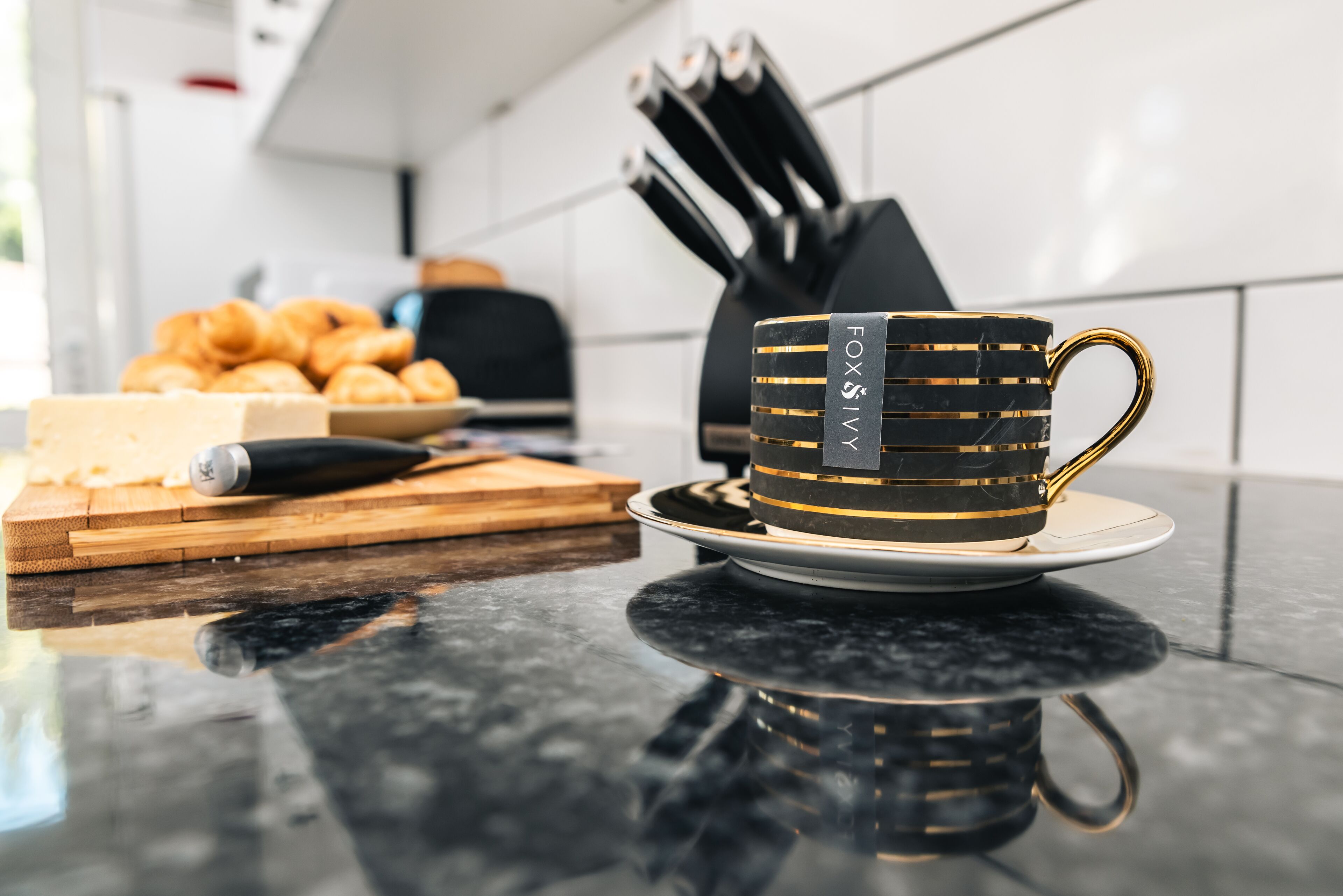 A kitchen counter with a black and gold coffee cup, a cutting board with cheese, and a knife block.