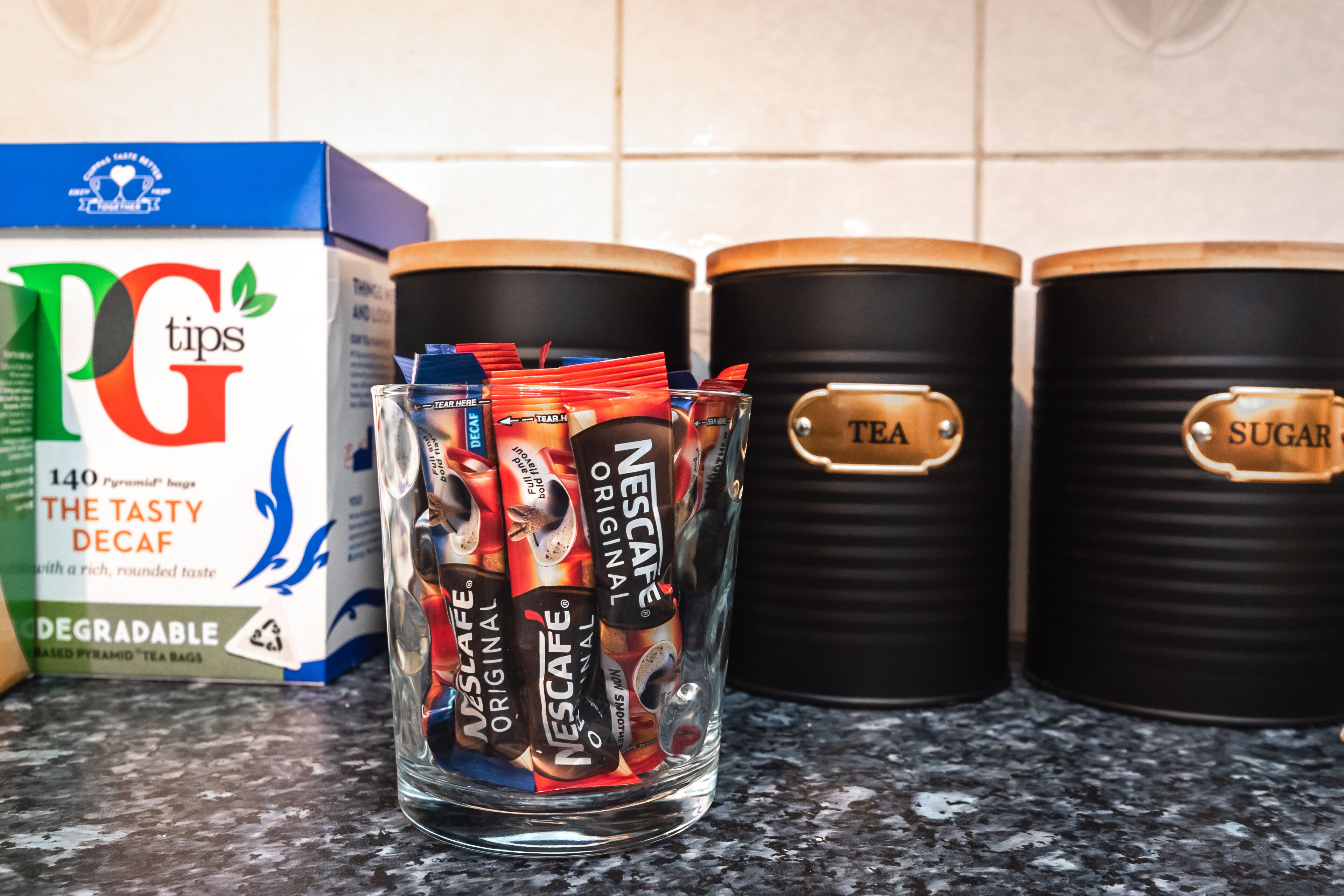 A kitchen counter with a box of PG tips tea bags, a glass jar filled with Nescafé Original tea packets, and two black containers labeled "TEA" and "SUGAR".