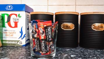 A kitchen counter with a box of PG tips tea bags, a glass jar filled with Nescafé Original tea packets, and two black containers labeled "TEA" and "SUGAR".