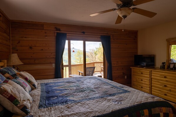 A wooden cabin bedroom with a bed, a dresser, a television, and a balcony with a view of trees.