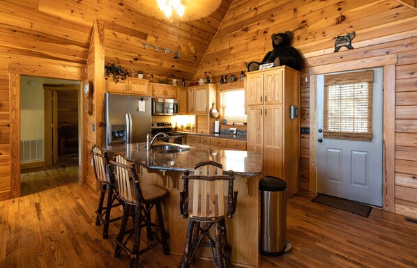 A wooden cabin interior with a kitchen featuring a refrigerator, microwave, stove, and bar stools.