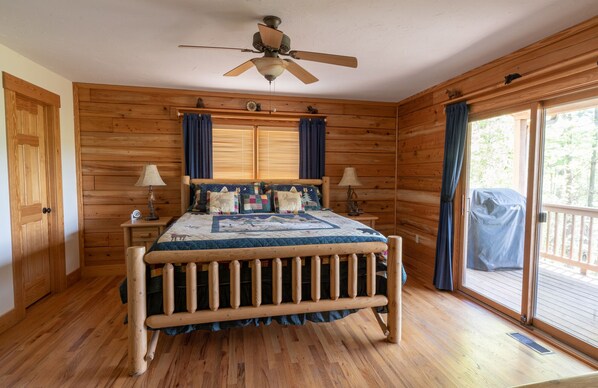 A wooden cabin bedroom with a bed, two bedside tables, a ceiling fan, and a large window with curtains.
