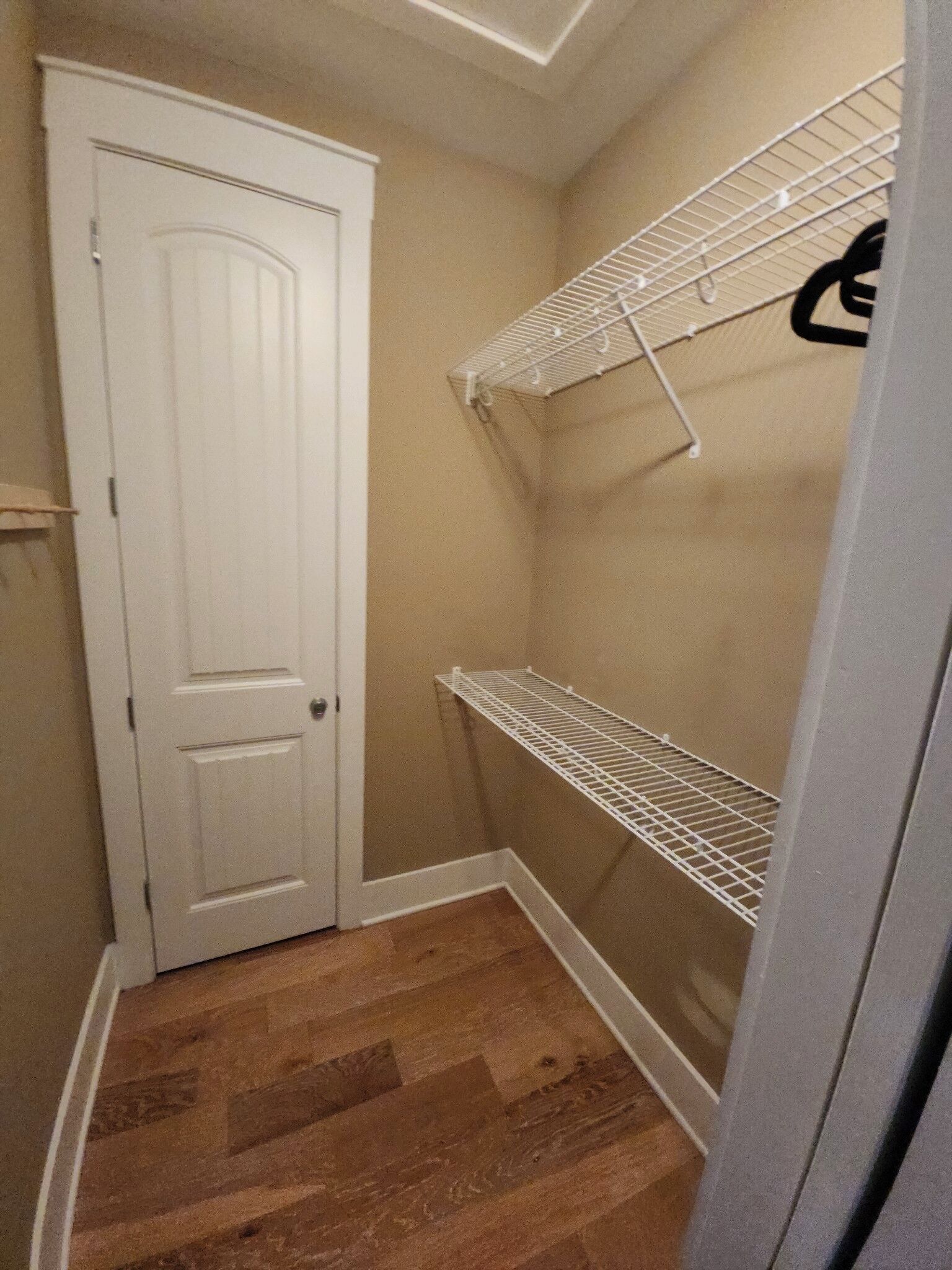 Un pasillo estrecho con una puerta blanca, suelo de madera y un estante metálico blanco adosado a la pared.