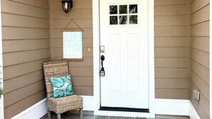 A front porch with a white door, a wicker chair with a cushion, a wall-mounted light fixture, and a welcome sign.