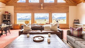 Un salon chaleureux avec une grande fenêtre donnant sur un paysage montagneux, un canapé en cuir et un coin repas avec une table en bois.