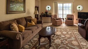 Un salon avec un canapé marron, deux fauteuils, une table basse et un téléviseur. Il y a deux fenêtres avec des rideaux, une horloge et un tableau encadré au mur.