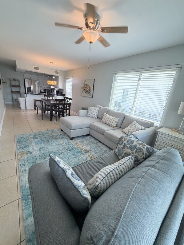 A modern living room with a grey sectional sofa, a dining area with a table and chairs, a kitchen in the background, and a ceiling fan.