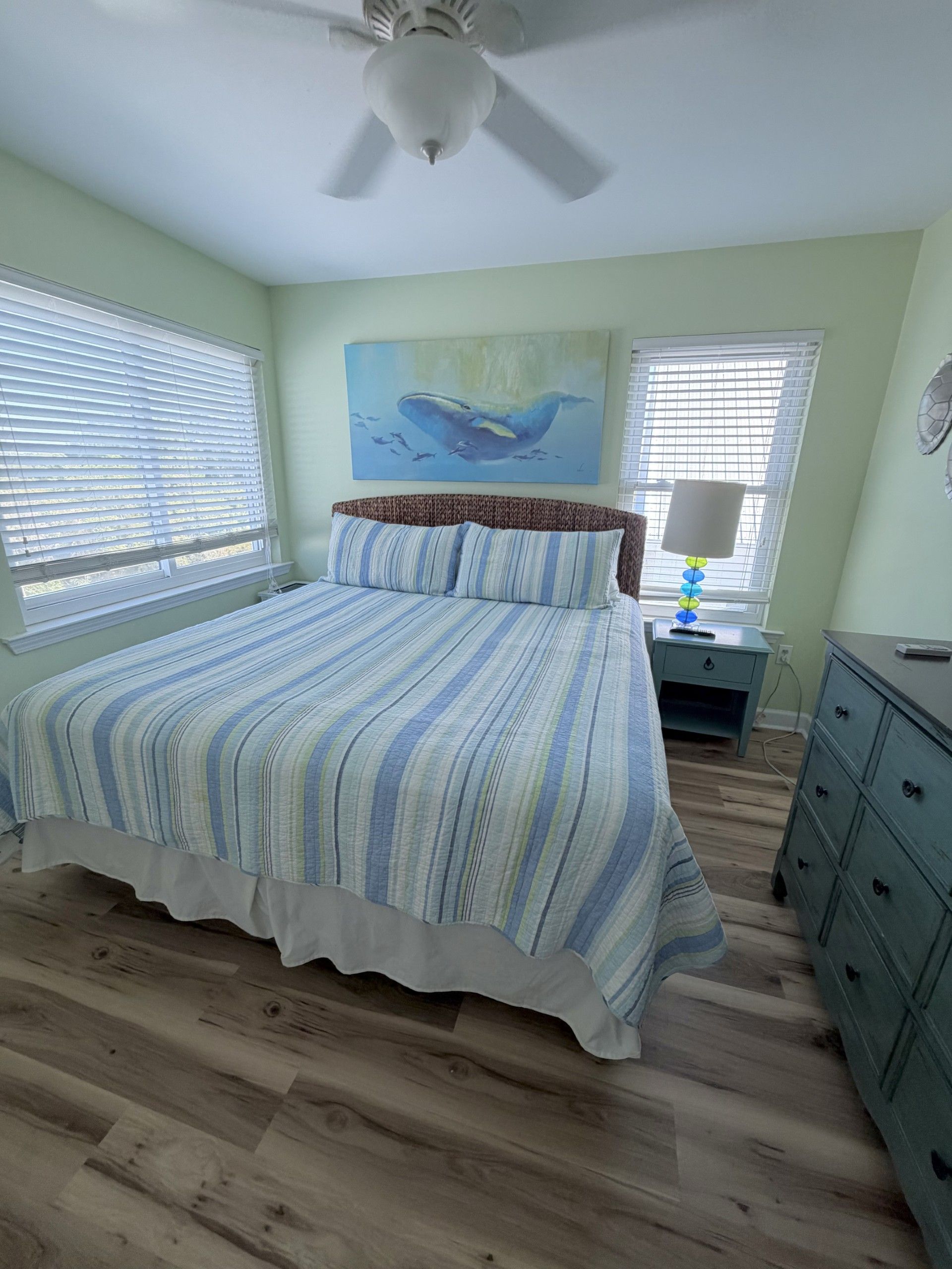Un dormitorio con cama, ventilador de techo, mesita de noche, cómoda y un cuadro en la pared.