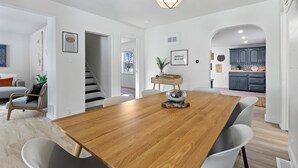 Un vaste espace de salle à manger avec une table en bois et des chaises modernes, un escalier menant à un autre niveau, et une cuisine dotée de meubles de couleur sombre.