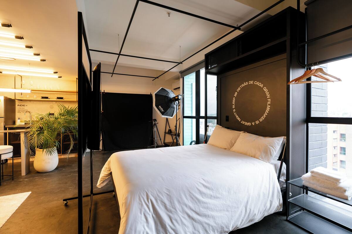 A modern hotel room with a large bed, a potted plant, a desk, and a view of the city through large windows.