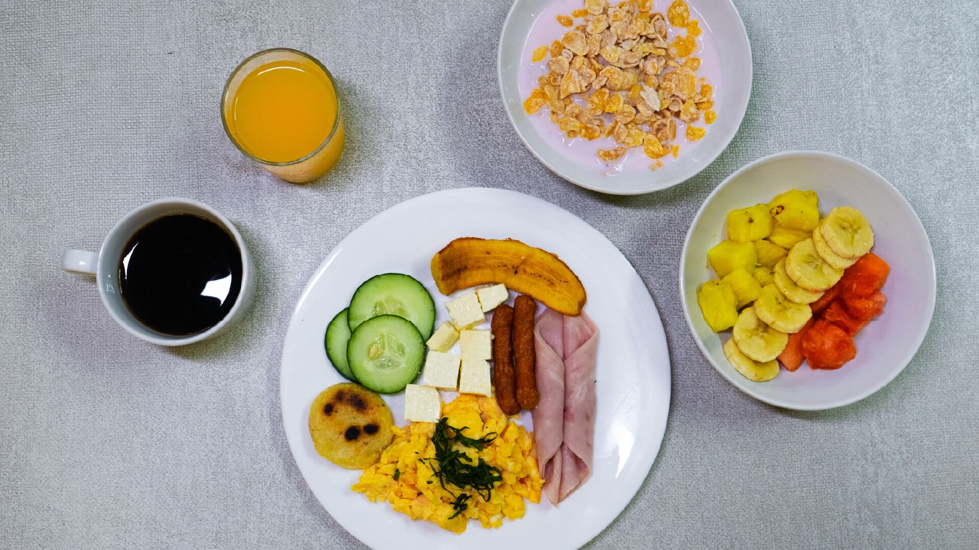 A breakfast plate with scrambled eggs, ham, cheese, cucumber slices, a banana, and a bowl of pineapple chunks.