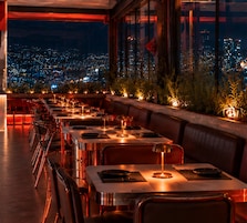 Un restaurante en la azotea con mesas y sillas dispuestas para comer, con vistas a un paisaje urbano nocturno.