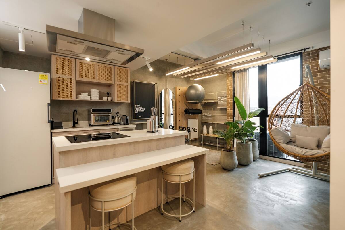 A modern kitchen with a central island, bar stools, a hanging chair, and a large window with a view.